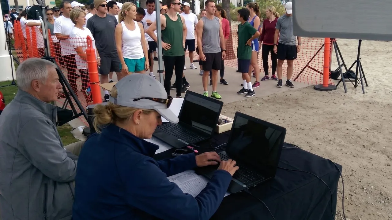 Two race officials working on laptops at a table while runners queue behind a barrier.