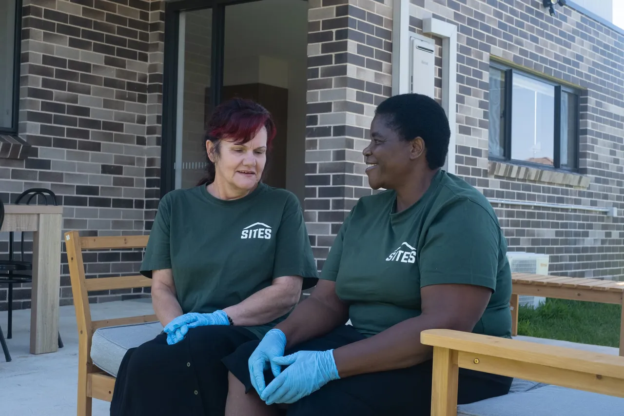 Two Sites support workers in green Sites T-shirts and blue gloves, talking on a bench outside a brick home.