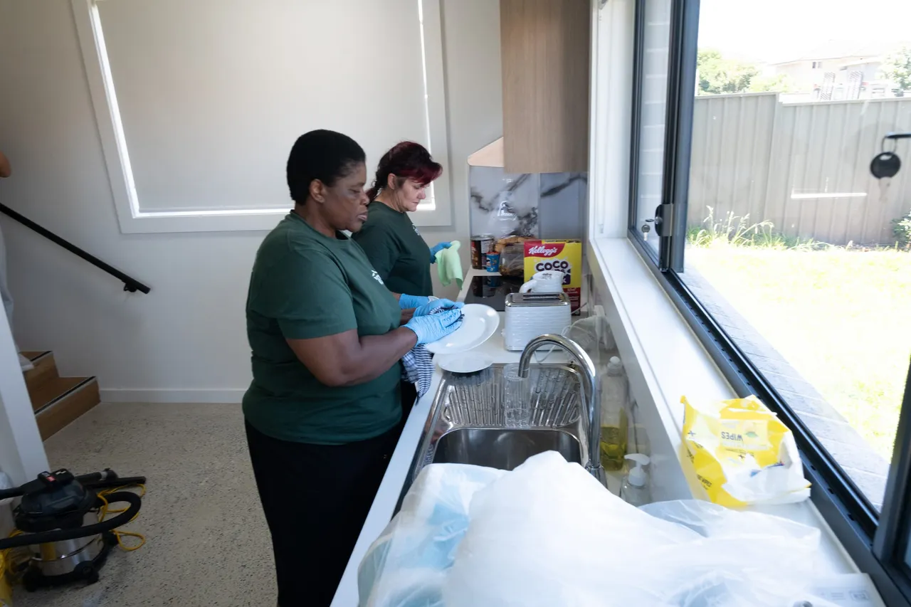 Two Sites support workers in green Sites T-shirts and blue gloves, washing and drying dishes at a kitchen sink.