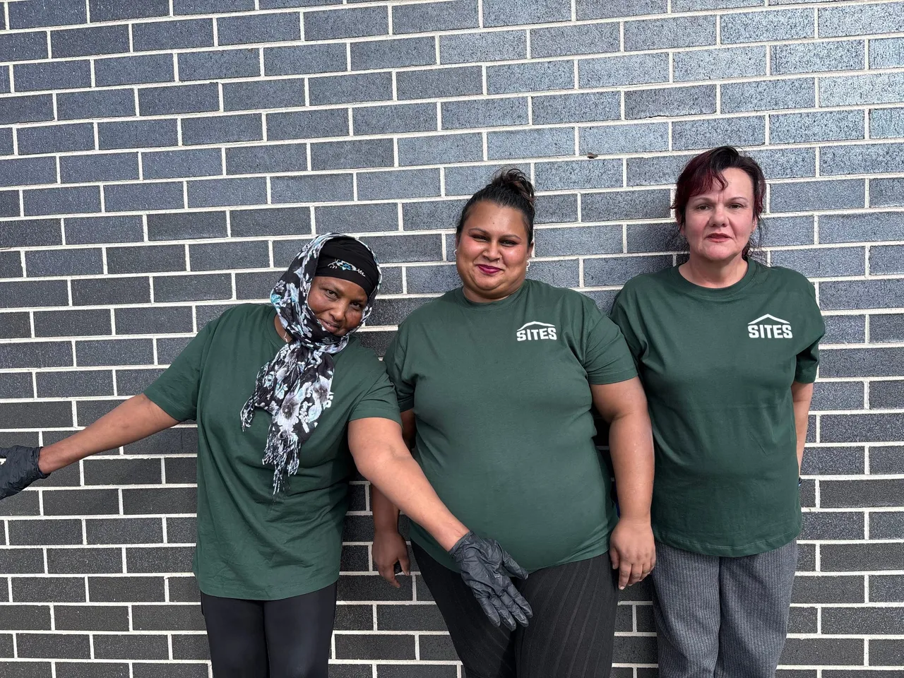 Three members of the Sites team in green Sites T-shirts, standing against a brick wall.