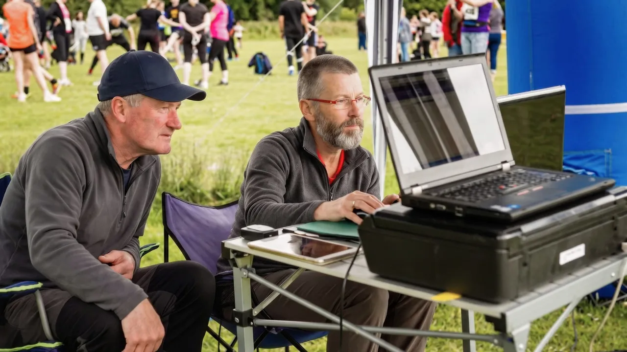 Two event officials watching a laptop at a timing desk, with runners warming up on the grass behind them.